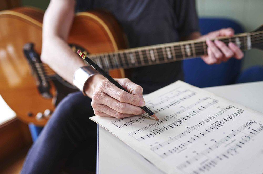 a person making notes on sheet music and holding a guitar.