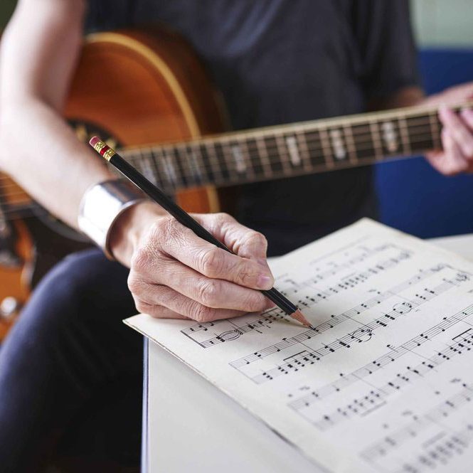 a person making notes on sheet music and holding a guitar.